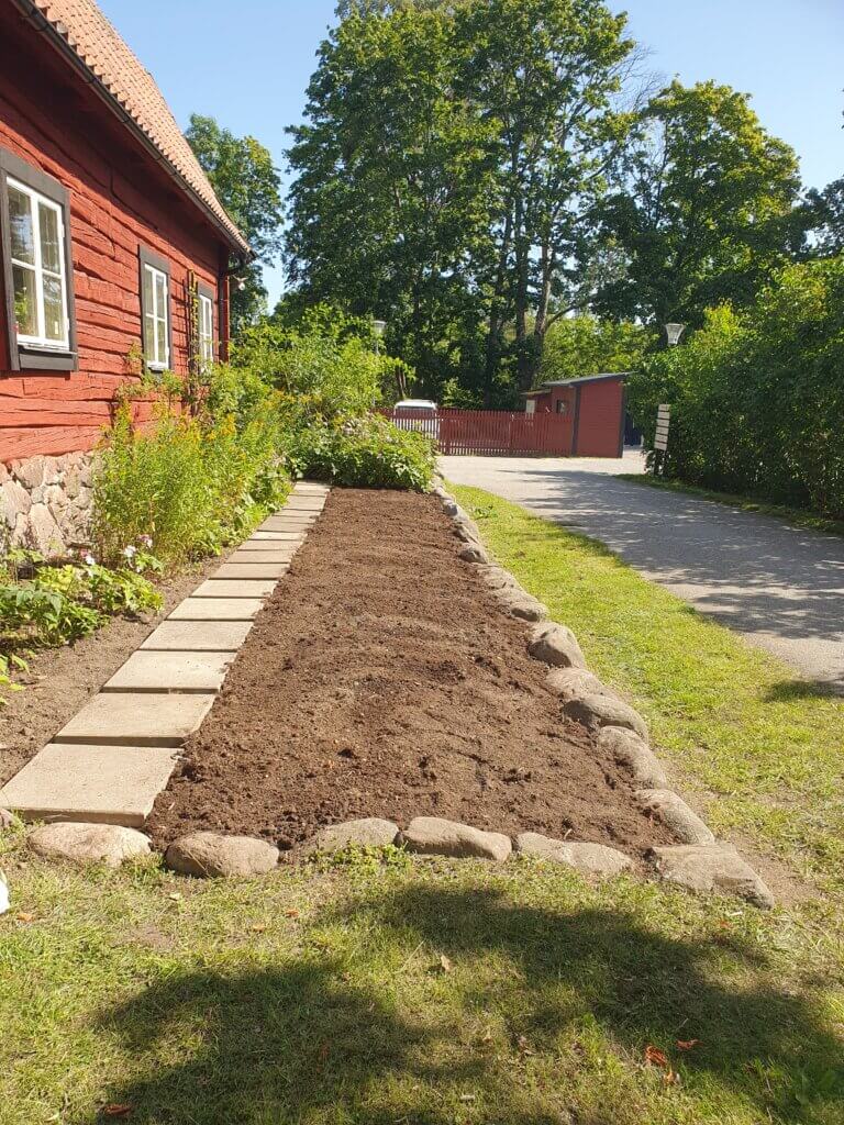 Nyanlagd rabatt med stenkanter vid rött hus.